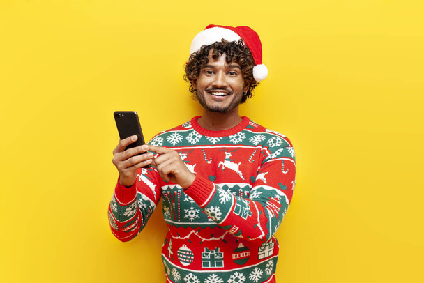 jonge krullende hindoe man in nieuwjaar kleding met behulp van smartphone op geel geïsoleerde achtergrond, hindoe man in santa hoed en kerst trui typen en selecteren van online - Foto, afbeelding