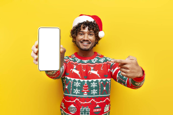νεαρός σγουρός Ινδουιστής άνδρας με ρούχα του νέου έτους που δείχνει λευκή οθόνη smartphone σε κίτρινο απομονωμένο φόντο, ο Ινδουιστής τύπος στο καπέλο santa και χριστουγεννιάτικο πουλόβερ διαφήμιση κινητό οθόνη σε απευθείας σύνδεση - Φωτογραφία, εικόνα