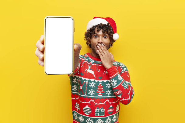 young shocked hindu man in new year clothes showing blank smartphone screen on yellow isolated background, surprised hindu guy in santa hat advertising mobile display online in amazement - Photo, Image