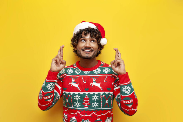 mladý indián muž v novém roce oblečení čeká a doufá s prsty zkříženými na žlutém izolovaném pozadí, hinduista chlap v Santa klobouk a vánoční svetr přání a sny - Fotografie, Obrázek