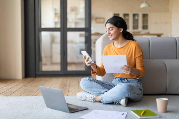 Молодая индийская женщина, работающая с ноутбуком и смартфоном дома, улыбающаяся фрилансер, сидящая на полу в гостиной, держащая бумаги и сообщения на мобильном телефоне, наслаждаясь удаленной работой - Фото, изображение