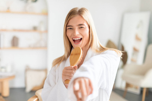 Positive woman in white robe singing with wooden brush, brushing her blonde hair in modern bathroom indoors, starting her day with positivity, enjoys grooming and carefree morning routine - Photo, Image