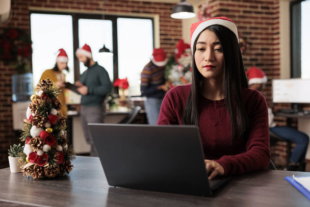 Asian woman corporate worker in santa hat working on project at winter seasonal holiday in decorated office. Company employee using typing on computer at festive coworking space - Photo, Image