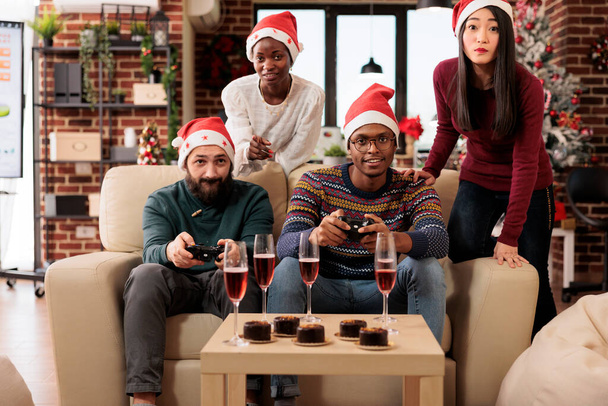 Ευτυχισμένοι συνάδελφοι ομάδα γιορτάζει τα Χριστούγεννα μαζί με το παιχνίδι βίντεο στο εορταστικό διακοσμημένο γραφείο. Συνάδελφοι με gamepads διασκεδάζοντας στο νέο έτος εταιρικό πάρτι στο χώρο εργασίας - Φωτογραφία, εικόνα