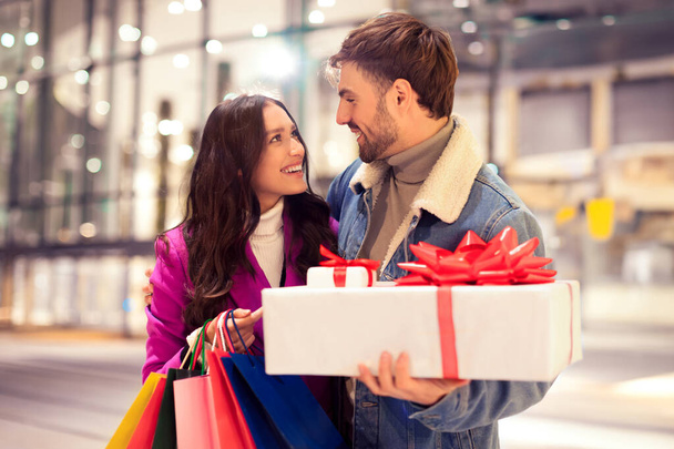 Ein fröhlicher Mann überrascht seine Freundin beim Weihnachtseinkauf mit einem verpackten Valentinstagsgeschenk und steht an einem gemütlichen Winterabend vor einem modernen Einkaufszentrum. Saisonale Verkäufe und Mode - Foto, Bild