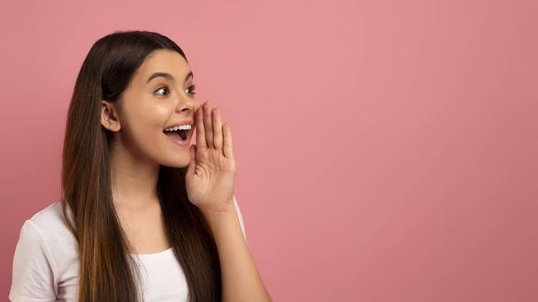 Excited teen girl with her hand near mouth shouting at copy space, happy female teenager sharing secret or making announcement, advertising offer, standing against pink background, panorama - Photo, Image