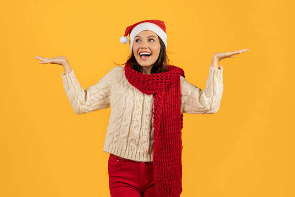 Kırmızı Noel Baba şapkalı ve atkılı neşeli genç bir kadın kollarını sanki görünmez nesneler taşıyormuş gibi açıyor. Bayram karşılaştırma reklamları için mükemmel. Stüdyo çekimi sarı arka planda. - Fotoğraf, Görsel