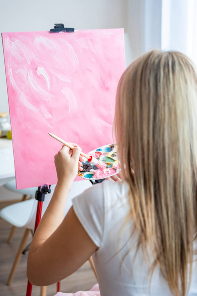 Вид ззаду молода жінка художник з палітрою і пензлем сидить і малює абстрактну рожеву картину на полотні біля вікна вдома. Концепція мистецтва та творчості. Високоякісна фотографія - Фото, зображення