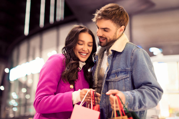 Cheerful shopaholics couple embracing standing with shopping bags, enjoying winter evening of holiday sales outside of store, perfect for Xmas seasonal shopping concepts, city mall background - Photo, Image
