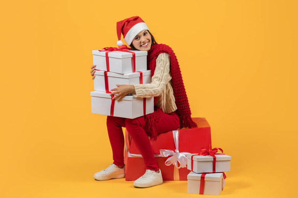 Χαρούμενη κυρία σε κόκκινο κρατώντας πολλά δώρα Χριστούγεννα κάθεται σε μεγάλο τυλιγμένο κουτί δώρου, γιορτάζει χαρούμενα, ιδανικό για εποχιακές διαφημίσεις αγορών και υπηρεσιών παράδοσης, στο στούντιο σε κίτρινο φόντο - Φωτογραφία, εικόνα