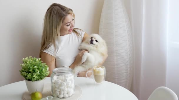 Güzel genç bir kadın Pomeranya köpeğiyle eğleniyor ve ev mutfağında kahve içiyor. Yüksek kalite 4k görüntü - Video, Çekim