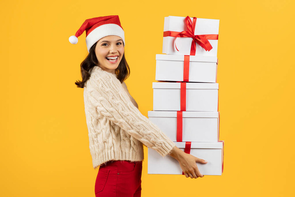 Szczęśliwa młoda dama uśmiechnięta stosem zapakowanych prezentów świątecznych, patrząca w kamerę, pozująca z zapakowanymi pudełkami prezentów w żółtym studio. Celebracja i koncepcja dawania prezentów - Zdjęcie, obraz
