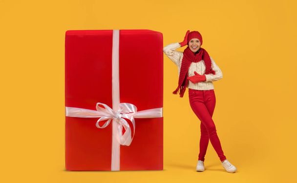 Ευτυχισμένη νεαρή κοπέλα με κόκκινο πλεκτό καπέλο ακουμπισμένη στο γιγάντιο τυλιγμένο χριστουγεννιάτικο δώρο ακτινοβολούν με χαμόγελο, στούντιο πυροβόλησε σε κίτρινο φόντο, πλήρους μήκους. Χειμερινή εορταστική περίοδο πωλήσεων - Φωτογραφία, εικόνα