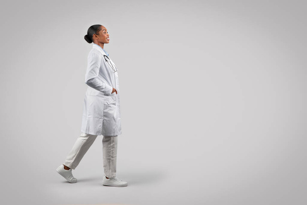 Pozitivní sebevědomá černá mladá dáma doktor v bílém kabátě, jít, izolované na šedém pozadí studia, plná délka. Lékařská péče, profesionální vyšetření a rychlá pomoc, práce - Fotografie, Obrázek