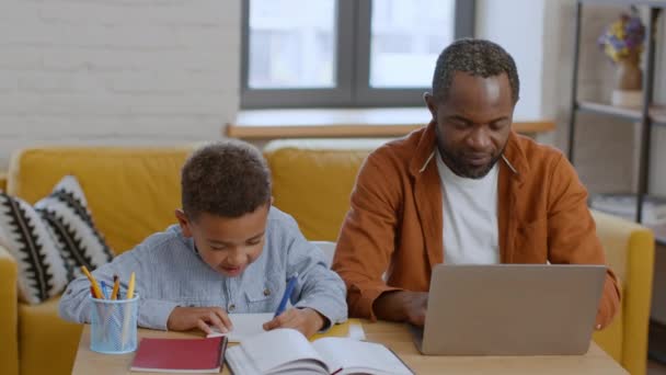 Trabaja para todos. Positivo afroamericano padre e hijo pasar tiempo útil en casa y sonriendo el uno al otro, chico haciendo la tarea, hombre trabajando en el ordenador portátil, espacio libre - Metraje, vídeo