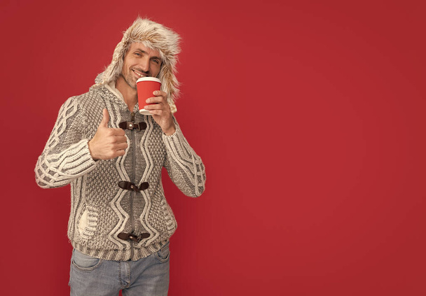 smiling male fashion model hold cup. winter fashion. morning coffee in take away cup. thumb up. grizzled guy in sweater and hat on red background. hoary man drinking coffee. - Foto, immagini