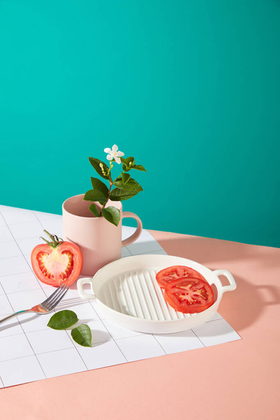 Verse tomatenschijfjes zijn gedecoreerd in een witte keramische plaat, ernaast staat een halve tomaat, een metalen lepel, een keramische kop en een camellia steel op een roze en turquoise achtergrond. Ruimte voor weergave. - Foto, afbeelding