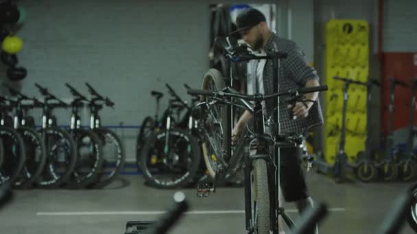 Tiro de mano de un joven mecánico caminando para montar en bicicleta en el puesto de trabajo, tomando asiento y aplicando lubricante a la cadena durante la jornada laboral en el garaje - Imágenes, Vídeo