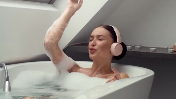 El metraje en cámara lenta 4K captura a una mujer feliz en un baño espumoso, vistiendo auriculares, perdida en la música y cantando en voz alta. La escena irradia relajación, alegría y el poder terapéutico de la música - Metraje, vídeo