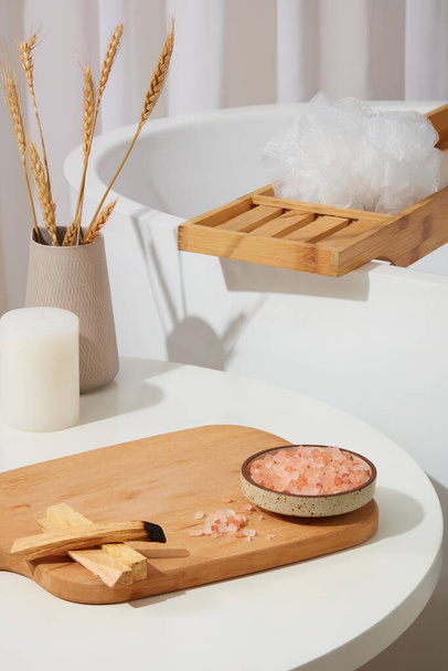 Деревянная доска с блюдом из розовой гималайской соли и дымом из древесины пало-санто. Ванная комната с ванной сзади. Пустое место для презентации продукта - Фото, изображение