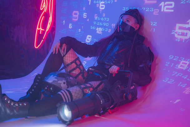 Una figura femminile, vestita in un futuristico ensemble tattico nero, si adagia a terra con un proiettore di grandi dimensioni su uno sfondo di simboli digitali proiettati - Foto, immagini