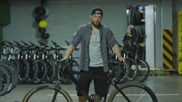 Közepes távolságból fiatal férfi áll kerékpárral a garázsban, és a kamerába néz - Felvétel, videó
