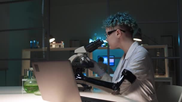 Inyección de arco medio de una joven técnica caucásica con un brazo protésico, con una bata blanca y guantes trabajando sola en el laboratorio de química - mirando la muestra bajo el microscopio, luego escribiendo en la computadora portátil - Metraje, vídeo