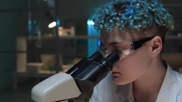 青いカーリーの髪を持つ若い女性の白人研究者の顔のクローズアップショット,薄暗く点灯した生化学実験室で単独で作業し,顕微鏡を調べながら実験を行っている - 映像、動画