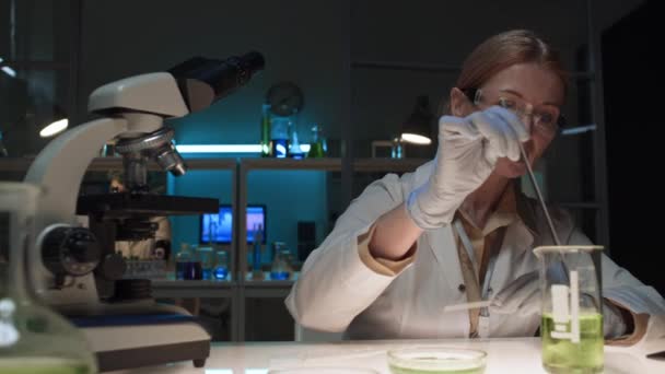 Средний снимок женщины-химика средних лет, работающей в биохимической лаборатории - помешивание зеленой жидкости во фляжке, подбор ее с пипеткой, аккуратное падение в чашку Петри и наблюдение реакции - Кадры, видео