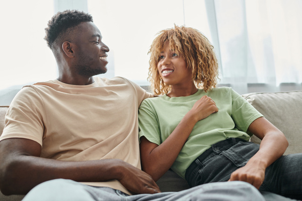 felice donna africana americana in bretelle guardando fidanzato mentre seduto sul divano, tempo di qualità - Foto, immagini