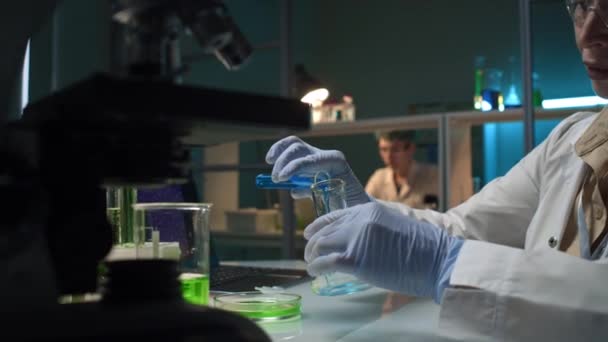 Střední detailní záběr rukou výzkumnice nalévající modrý roztok z injekční lahvičky do baňky, přidávající zelenou tekutinu, pak míchající obsah v kruhovém pohybu a ověřující výsledek - Záběry, video