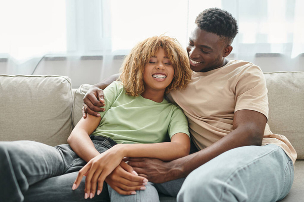 veselý černošky muž objímající šťastný přítelkyně v rovnátka zatímco sedí na útulný gauč v obývacím pokoji - Fotografie, Obrázek