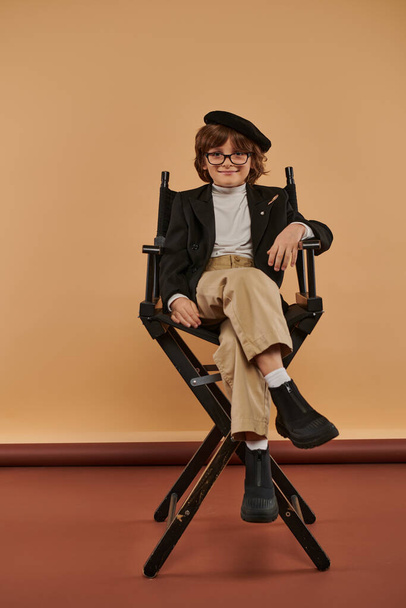 niño feliz en ropa casual se sienta en silla de director sobre fondo beige, niño y profesión - Foto, Imagen