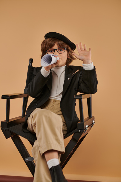 elegante chico en boina se sienta en silla de director, hablando en un pedazo de papel laminado y mostrando cinco - Foto, imagen