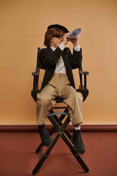 χαριτωμένο αγόρι με μοντέρνα ρούχα κάθεται στην καρέκλα διευθυντής και κοιτάζοντας μέσα από την τρύπα στο χαρτί έλασης - Φωτογραφία, εικόνα