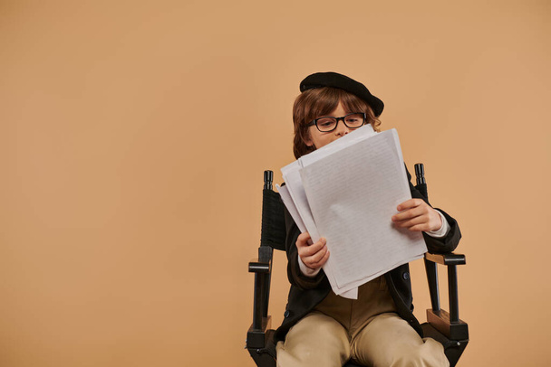 zajęty chłopiec w okularach siedzi w fotelu reżysera, patrząc na papiery i pochłonięty czytaniem - Zdjęcie, obraz