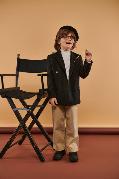 εμπνευσμένο αγόρι που στέκεται κοντά σε καρέκλα σκηνοθέτη, με ένα χαμόγελο στο πρόσωπο που δείχνει σημάδι ιδέα, επάγγελμα - Φωτογραφία, εικόνα