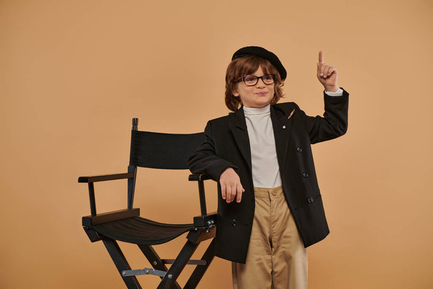 niño feliz de pie cerca de la silla del director, con una sonrisa en la cara que muestra signo de idea, profesión - Foto, Imagen