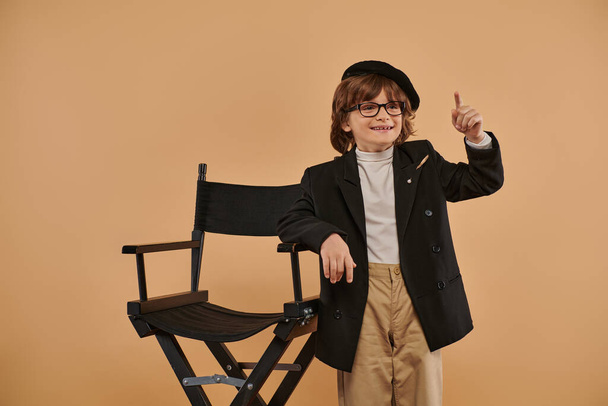 alegre chico de pie cerca de director silla, con una sonrisa en la cara mostrando idea signo, profesión - Foto, imagen