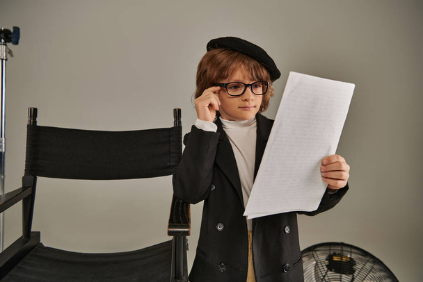 chico con estilo en gafas y boina guion de lectura en papel sobre gris, niño como director de cineasta - Foto, Imagen