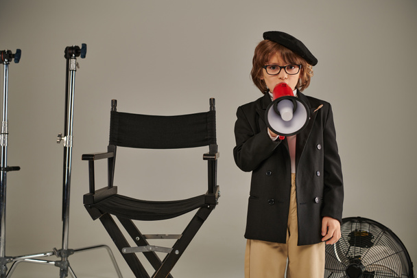 αγόρι ως σκηνοθέτης στέκεται με αυτοπεποίθηση κοντά καρέκλα σκηνοθέτη και μιλώντας σε μεγάφωνο, γκρι φόντο - Φωτογραφία, εικόνα
