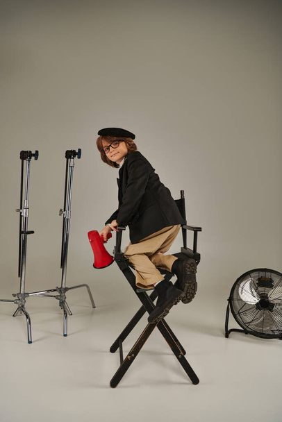 Yönetmen çocuk yönetmen koltuğuna tırmanıyor ve elinde gri fonda kırmızı megafon tutuyor. - Fotoğraf, Görsel