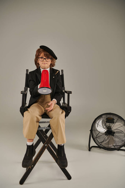 αυτοπεποίθηση αγόρι κινηματογραφιστής σε μπερέ και γυαλιά κάθεται σε καρέκλα σκηνοθέτη και κρατώντας κόκκινο μεγάφωνο - Φωτογραφία, εικόνα