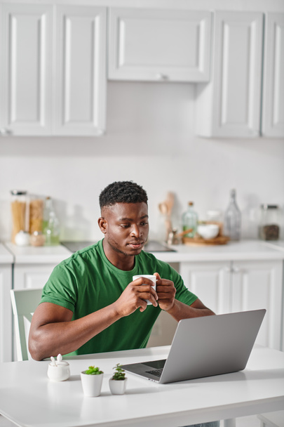homme afro-américain profiter d'une tasse de café tout en utilisant un ordinateur portable dans la cuisine, pigiste à la maison - Photo, image