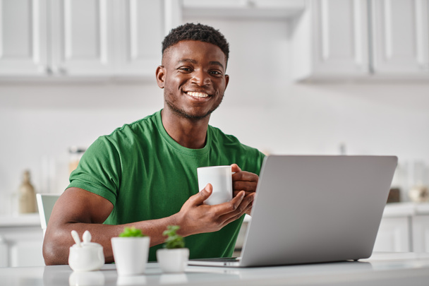 cheerful black man enjoying cup of coffee while using laptop in kitchen, freelancer at home - Photo, Image