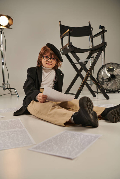 παιδί με γυαλιά και μπερέ διαβάζει σενάριο και κάθεται στο πάτωμα, αγόρι ως σκηνοθέτης - Φωτογραφία, εικόνα