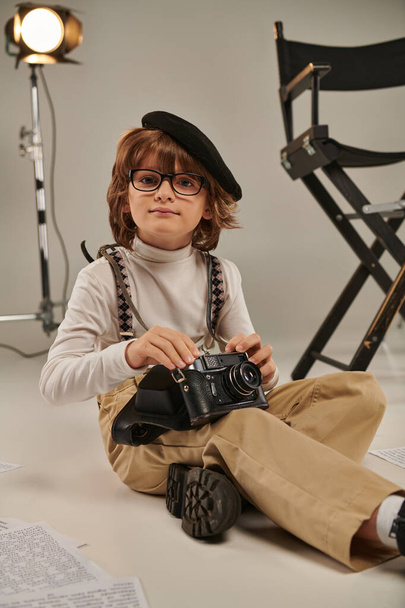 мальчик в берете держит ретро-камеру и сидит на полу возле режиссерского кресла, молодой фотограф - Фото, изображение