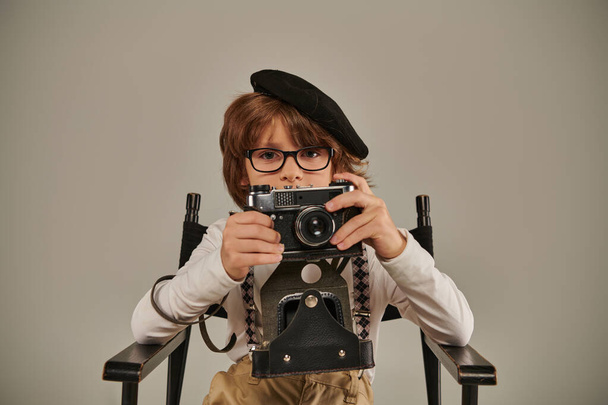chłopiec w berecie i okulary trzymając zabytkowy aparat podczas siedzenia na fotelu reżysera, młody fotograf - Zdjęcie, obraz