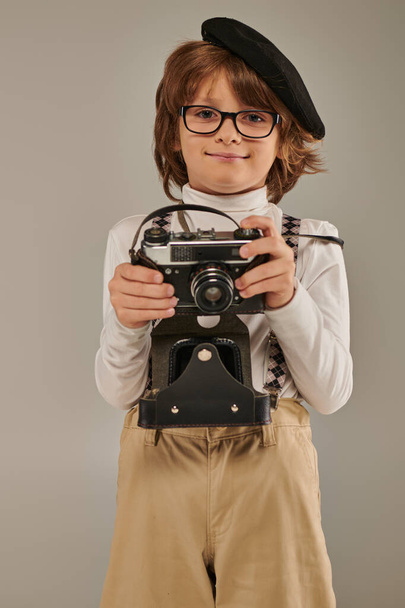 happy boy, young photographer in beret and suspenders holding retro camera while standing in studio - Фото, зображення