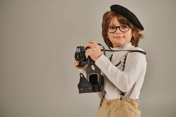 pleased boy, young photographer in beret and suspenders taking photo on retro camera in studio - Photo, Image